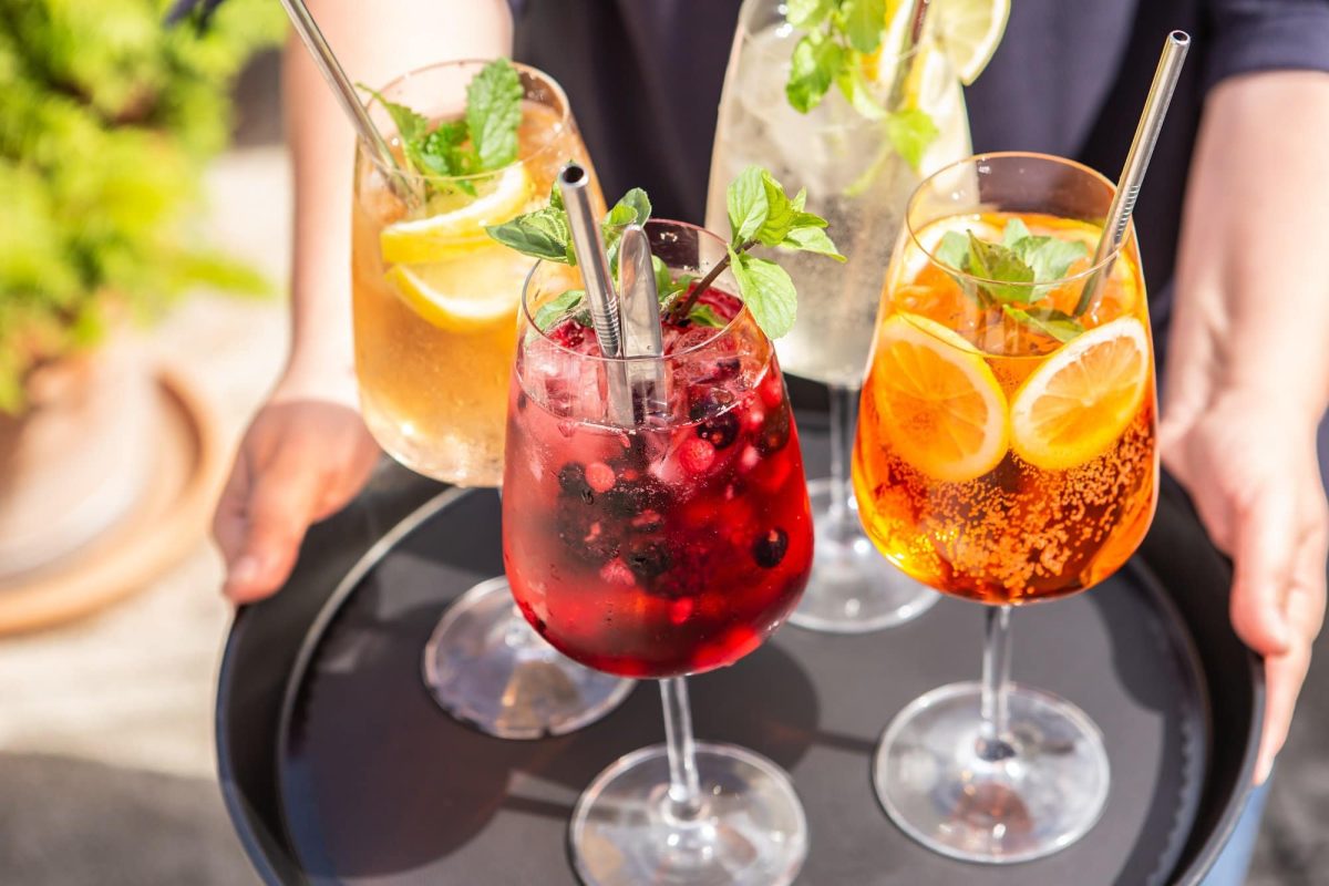 4 verschiedene Spritz-Cocktails auf einem Tablett, das von einer Frau getragen wird. Man sieht nur ihre Hände.