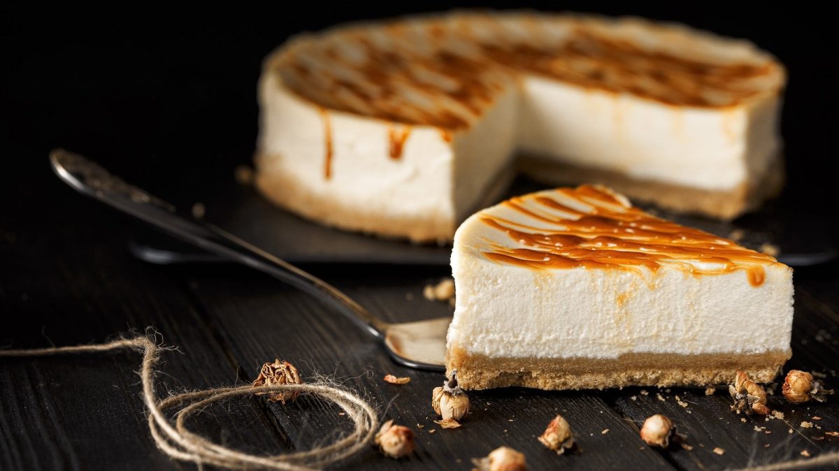 Ein Stück Tahini-Cheesecake mit Salzkaramellsauce, dahinter sieht man unscharf den angeschnittenen Kuchen.