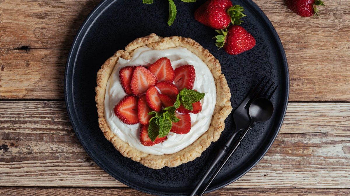 Gefüllte Tarte mit Vanillecreme und marinierten Erdbeeren mit Minzblättern und Erdbeeren auf schwarzem Teller auf Holz. Daneben schwarzes Besteck. Draufsicht.