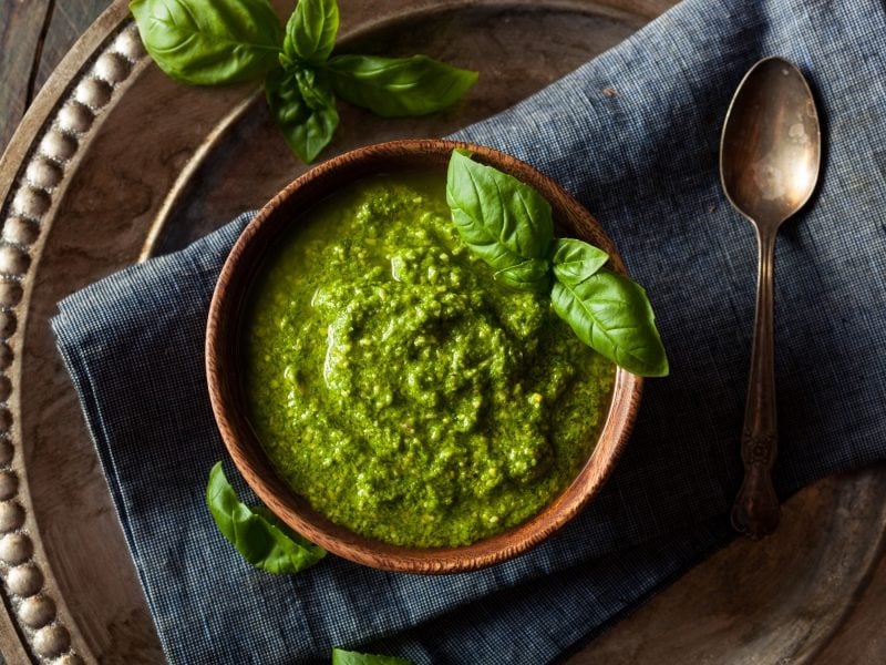 Cremiges Pesto verde garniert mit Basilikumblättern, serviert in Holzgefäß auf gewebter Tischdecke.