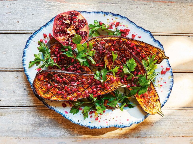 Vegan vom Grill: gegrillte Aubergine mit Tamarinde, Granatapfelkernen und frischer Petersilie auf einem Teller in der Draufsicht.