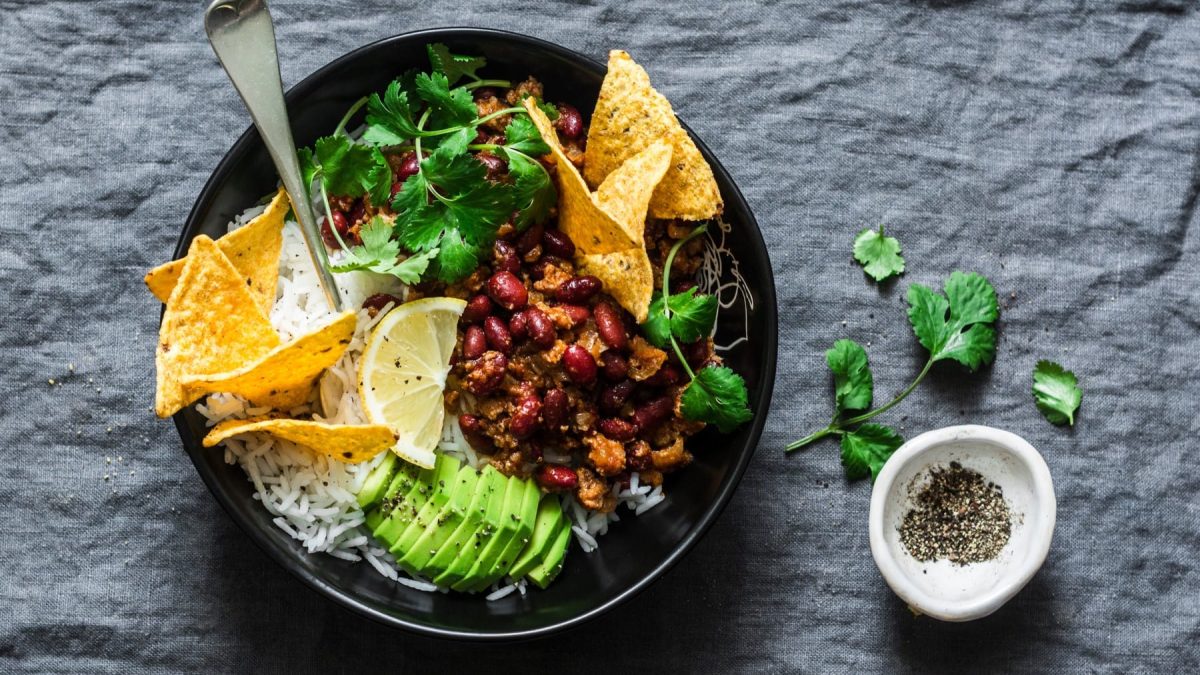 Draufsicht: Vegane Burrito-Bowl mit Tortilla-Chips und einer Gabel in einer schwarzen Schüssel auf grauem Stoffuntergrund, daneben eine Schale schwarzer Pfeffer und etwas Koriander.