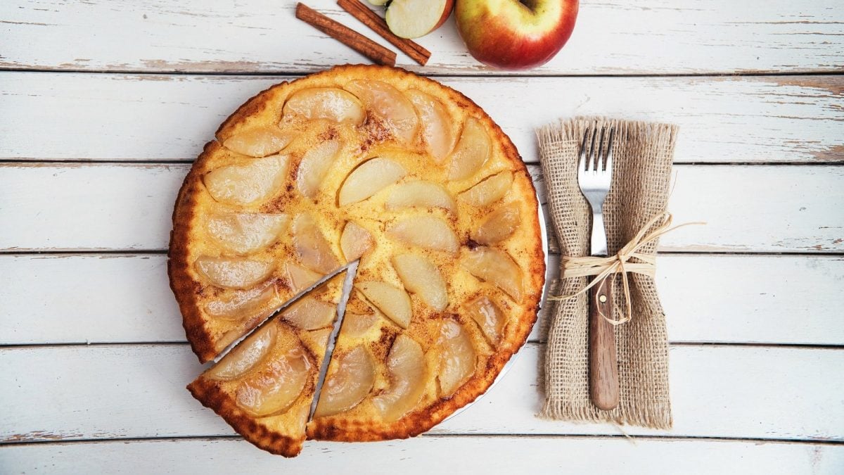 Ein angeschnittener veganer Apfelkuchen in der Draufsicht, daneben eine Gabel und frische Äpfel.