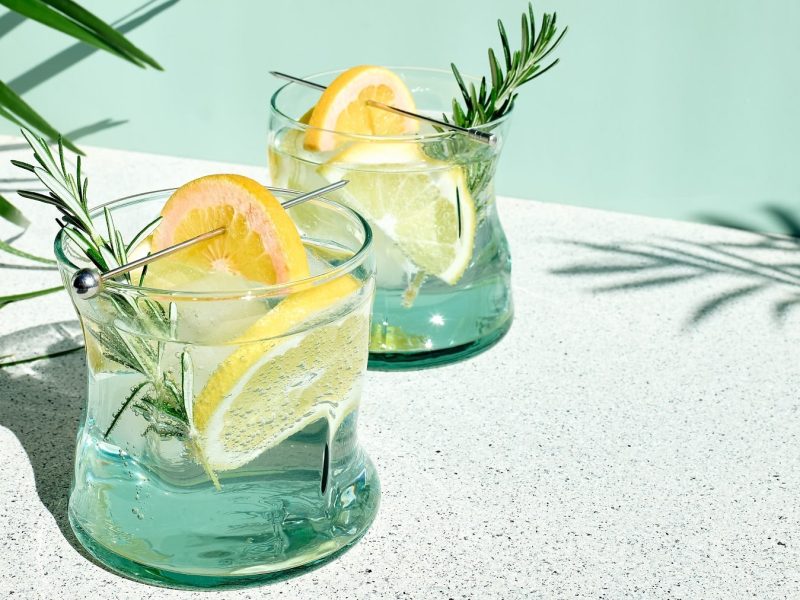 Zwei Gläser mit dem Vodka Tonic auf einem hellen Steintisch mit grünem Hintergrund und Palmen links und rechts.
