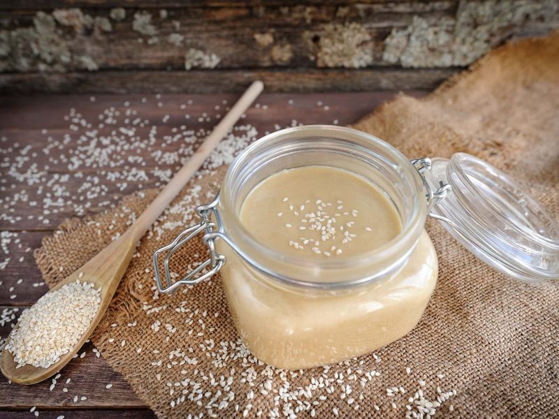 Draufsicht ein Twist-Off-Glas mit Tahin liegt auf einem Untergrund aus Holz. Daneben liegen verstreut helle Sesamsamen sowie ein Holzlöffel.