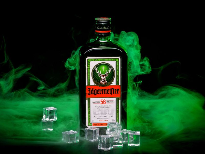 Welche Kräuter sind in Jägermeister? Eine Flasche Jägermeister mit Eiswürfeln vor schwarzem Hintergrund auf schwarzem Gruns. Grüner Nebel in Schwaden dynamisch im Bild. Frontalansicht.