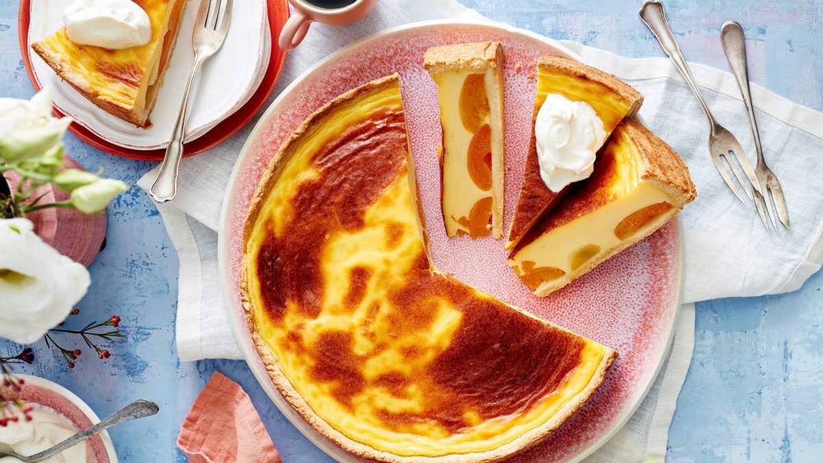Angeschnittener Aprikosen-Puddingkuchen mit Schlagsahne auf einem rosa Teller. Darum auf blauem Untergrund Geschirr und eine Pflanze.