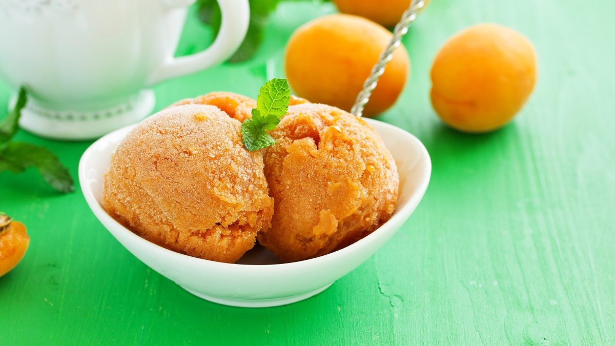 Drei Kugeln Aprikosen-Sorbet mit feiner Vanille ohne Eismaschine in weißer Schale mit Minzblatt auf grünem Untergrund. Im Bild eine weiße Kann und Aprikosen. Draufsicht.