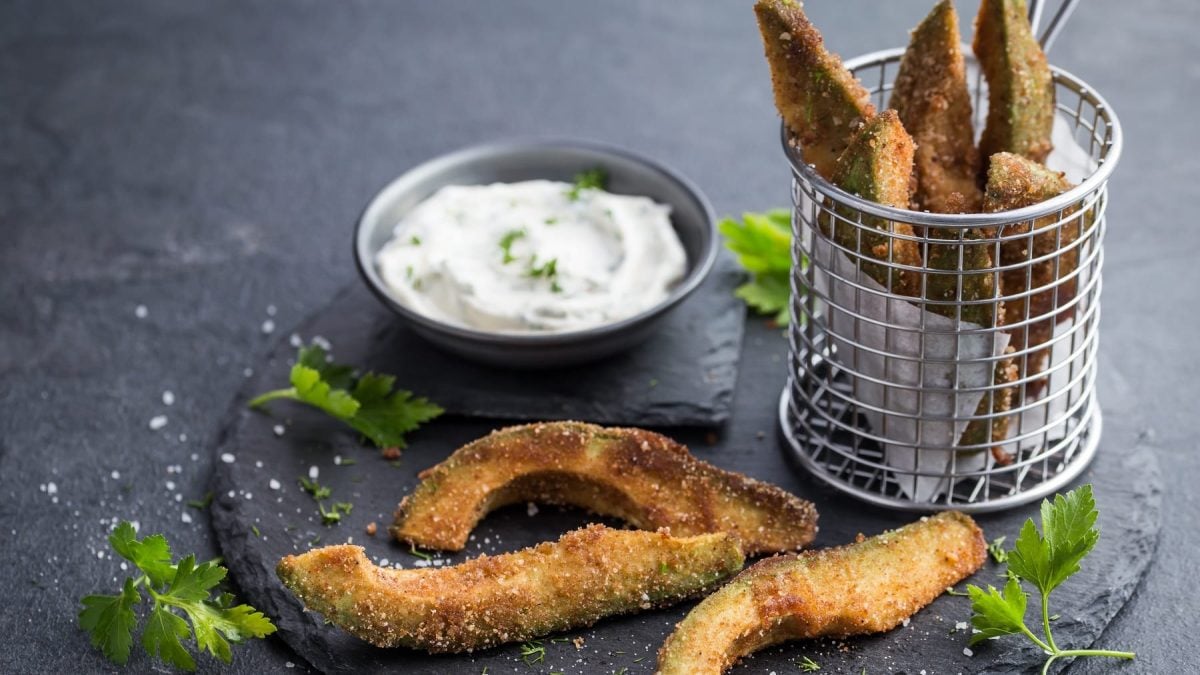 Drei Avocado-Fries auf einer runden Schiefertafel mit einem runden Frittierkorb aus Metall voller Avocado-Fries. Außerdem eine kleine Schale mit der weißen Sauce.