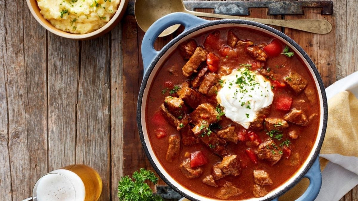 Ein Eintopf mit Gulasch auf einem Holztisch, darüber ein Suppenschöpfer. Links unten auf dem Tisch steht ein Bier und links oben ein Kartoffelpüree.