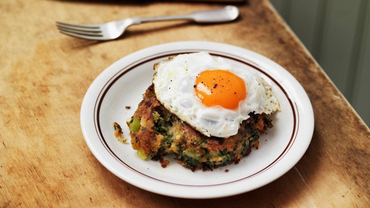 Draufsicht: Auf einem Holztisch steht eine weißer Runder Teller mit Bubble and Squeak mit einem Spiegelei. Daneben liegt eine Gabel.