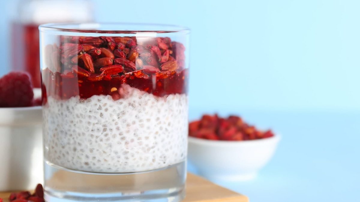 Ein kleines Glas mit dem Chia Pudding und darauf Himbeeren und Goji-Beeren auf der linken Seite des Bildes. Alles vor hellblauem Unter- und Hintergrund.