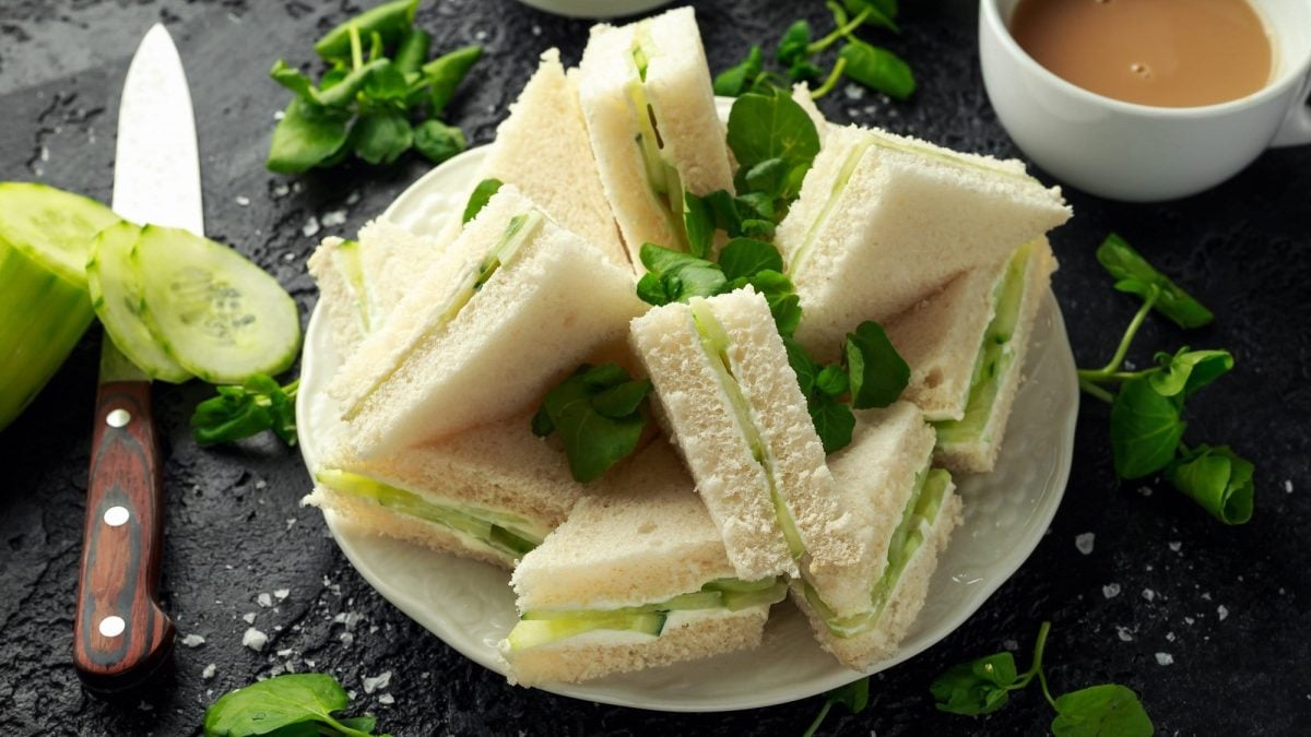 Draufsicht: dunkler Untergrund, ein Teller mit diagonal geschnittenen Cucumber Sandwiches. Daneben sind eine Tasse Kaffee und ein langes Küchenmesser.
