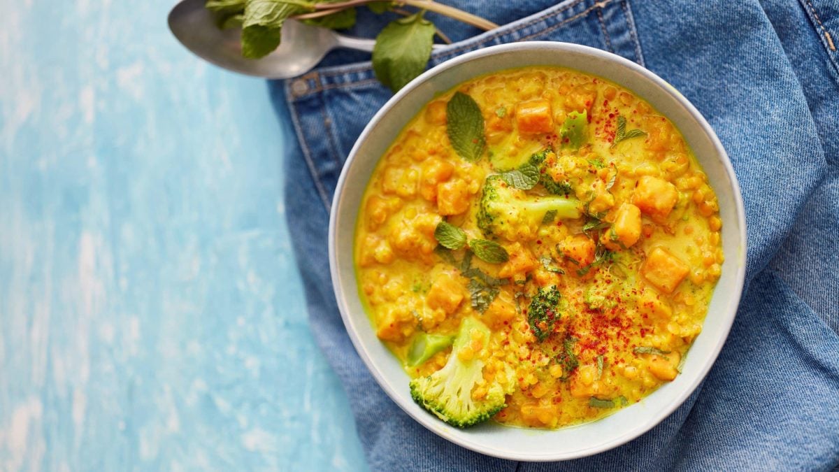 Eine hellgraue Bowl mit rotem Linsencurry auf einem Jeansstoff auf einem hellblauen Hintergrund. Oben ein Löffel und Minzblätter.