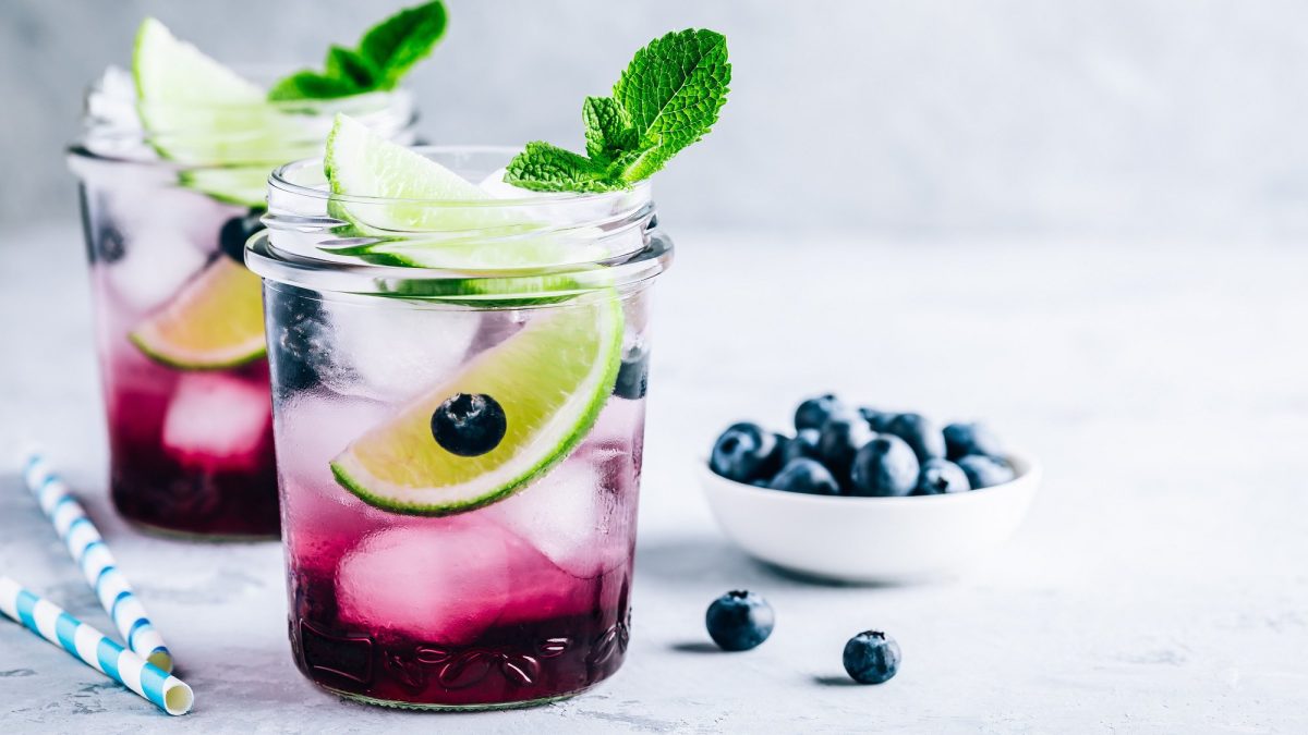 Zwei Gläser Eistee mit Blaubeeren und Minze, darin auch Limettenspalten und Eiswürfel, daneben Strohhalme und eine Schale Blaubeeren.