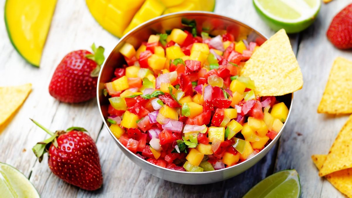 Eine Schüssel Erdbeer-Mango-Salsa mit Koriander, drumherum frische Zutaten und Nachos.