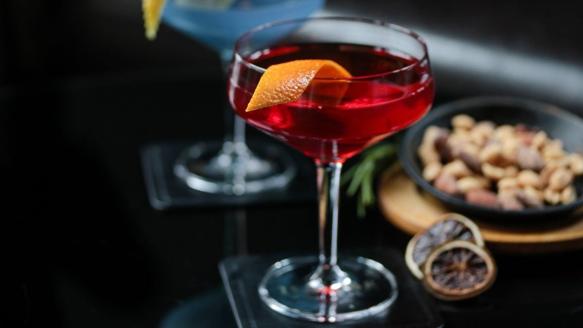 Frontal: ein Glas Gin Dubonnet auf einem dunklen Untergrund. Daneben steht eine Schale mit Nüssen.