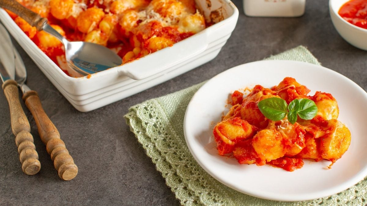 Ein weißer Teller mit dem Gnocchi-Ofengericht und der Auflaufform auf einem grauen Tisch mit Besteck und einer grünen Tischdecke.