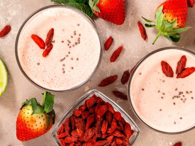 Zwei Gläser mit dem Shake von oben fotografiert. Daneben zahlreiche Zutaten des Shakes wie die Erdbeeren, Goji-Beeren und Limettenspalten.