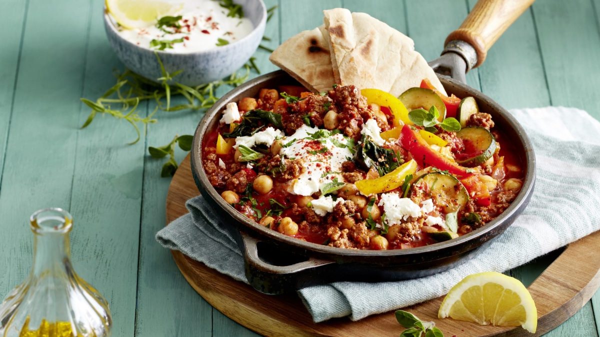 Eine Griechische Hack-Gemüse-Pfanne auf einem Geschirrtuch auf einem Holzbrett auf einem grünen Hintergrund. Links oben eine weiße Sauce in einer kleinen Schüssel, links unten eine Flasche mit Olivenöl.