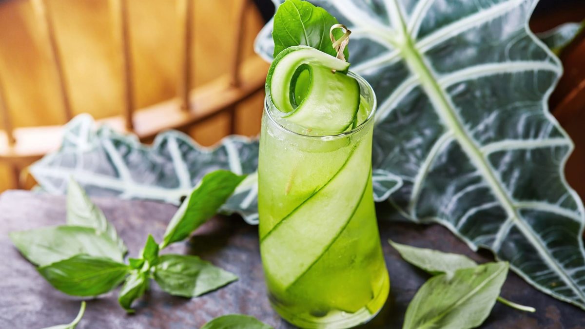 Ein Glas mit dem Gurkenwasser auf einem grauen Tisch. Im Hintergrund eine Holzfassade und eine Monstera-Pflanze.