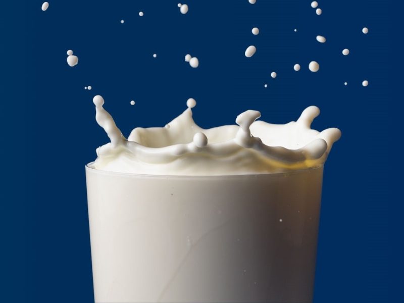Ein Tropfen fällt in ein randvolles Glas Milch, erzeugt eine spritzende Krone und verteilt feine Milchtröpfchen vor dunklem Hintergrund.
