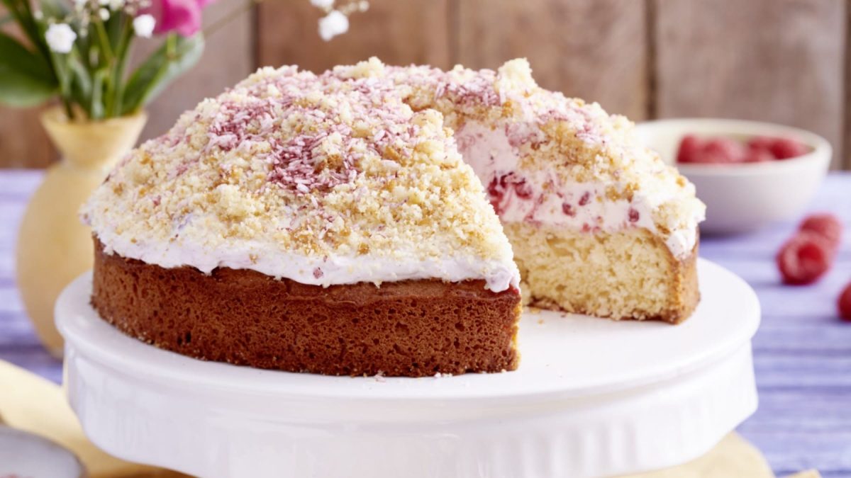 Ein Himbeer-Maulwurfskuchen auf einem weißen Tortenteller von vorne fotografiert auf einer violetten Fläche. Links ein Blumenstrauß, rechts eine Schüssel mit Himbeeren.