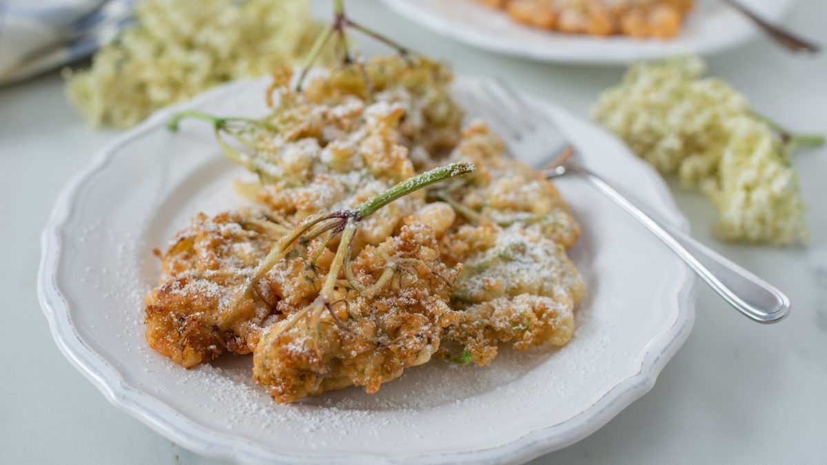 Holunderblüten frittieren Holunderblüten im Backteig auf weißem Teller auf weißem Untergrund. Daneben frische Holunderblüten, eine Gabel und ein weiterer Teller angedeutet. Draufsicht.