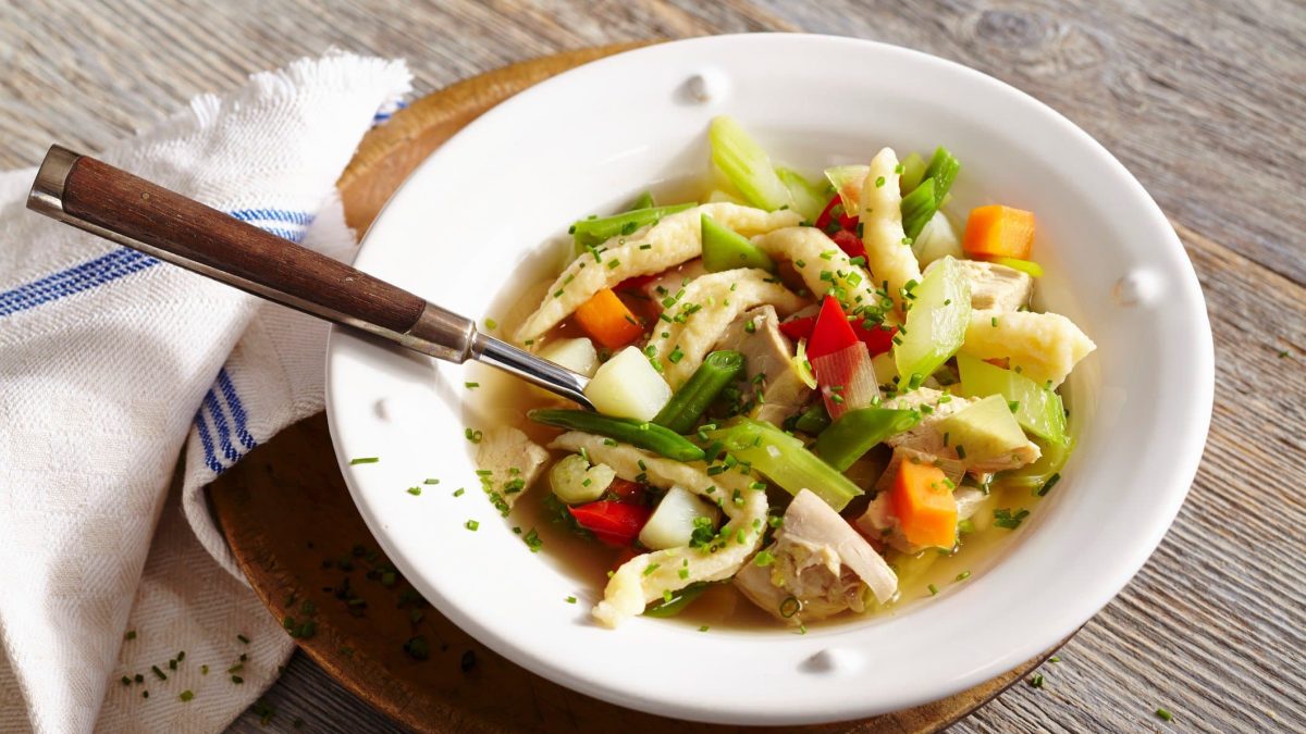Hühnersuppe mit Pizokel und Sommergemüse und Schnittlauch in weißem Suppenteller auf Holztisch. In der Suppe ein Suppenlöffel mit Holzgriff. Links ein blau-weißes Küchentuch. Draufsicht.