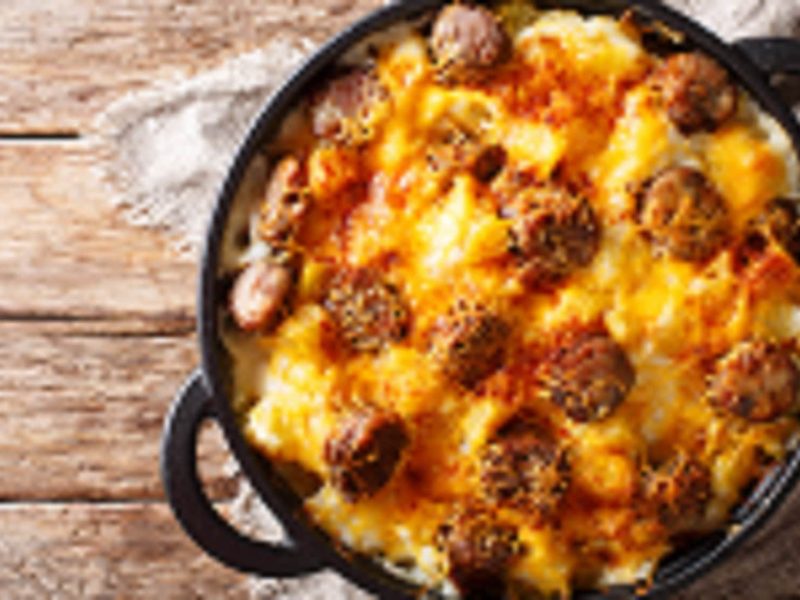 Draufsicht: eine Pfanne Jolean's Cheese Potato steht auf einem Untergrund aus Holz. es handelt sich um eine überbackene Würstchen-Pfanne mit Kartoffeln.