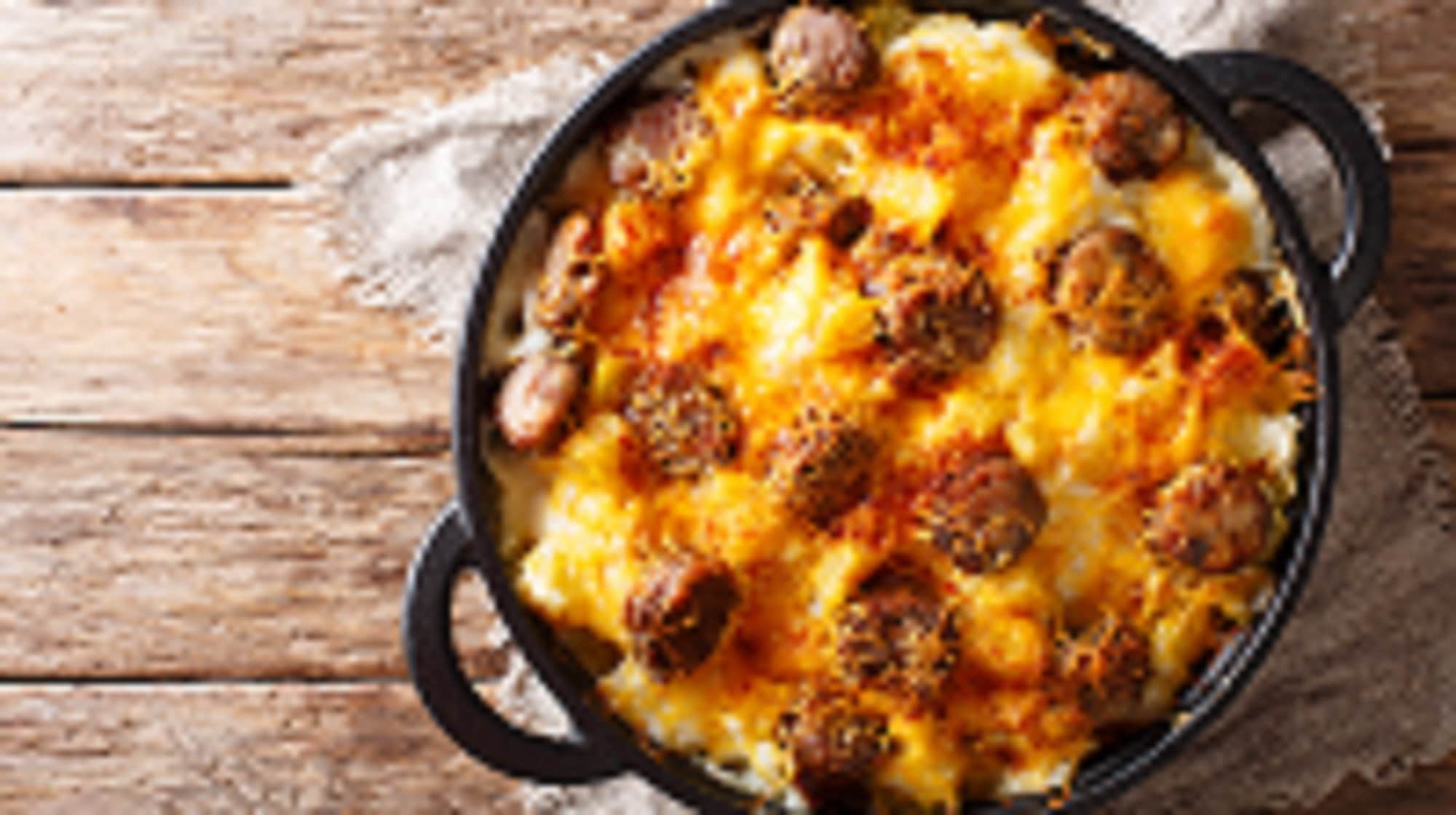 Draufsicht: eine Pfanne Jolean's Cheese Potato steht auf einem Untergrund aus Holz. es handelt sich um eine überbackene Würstchen-Pfanne mit Kartoffeln.