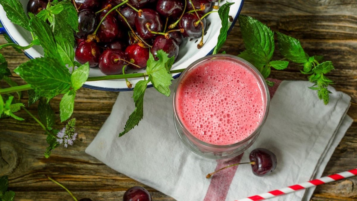 Kirsch-Smoothie mit Skyr und frischer Minze im Glas auf grauem Tuch und Holztisch. Daneben eine Schale mit Kirschen und Minzstängeln. Im Bild ein rot-weißer Strohhalm, eine Kirsche und Minzblätter. Draufsicht.
