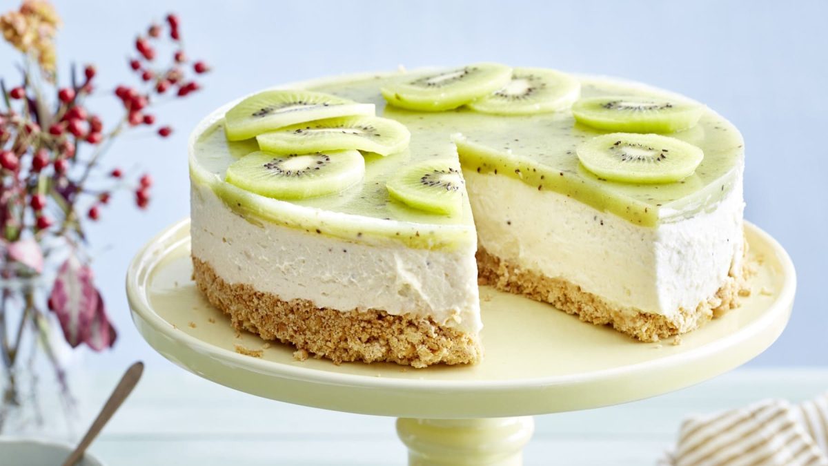 Ein Kiwi-Cheesecake auf einer hellgelben Tortenplatte vor einem bläulichen Hintergrund. Links ein Blumenstrauß.