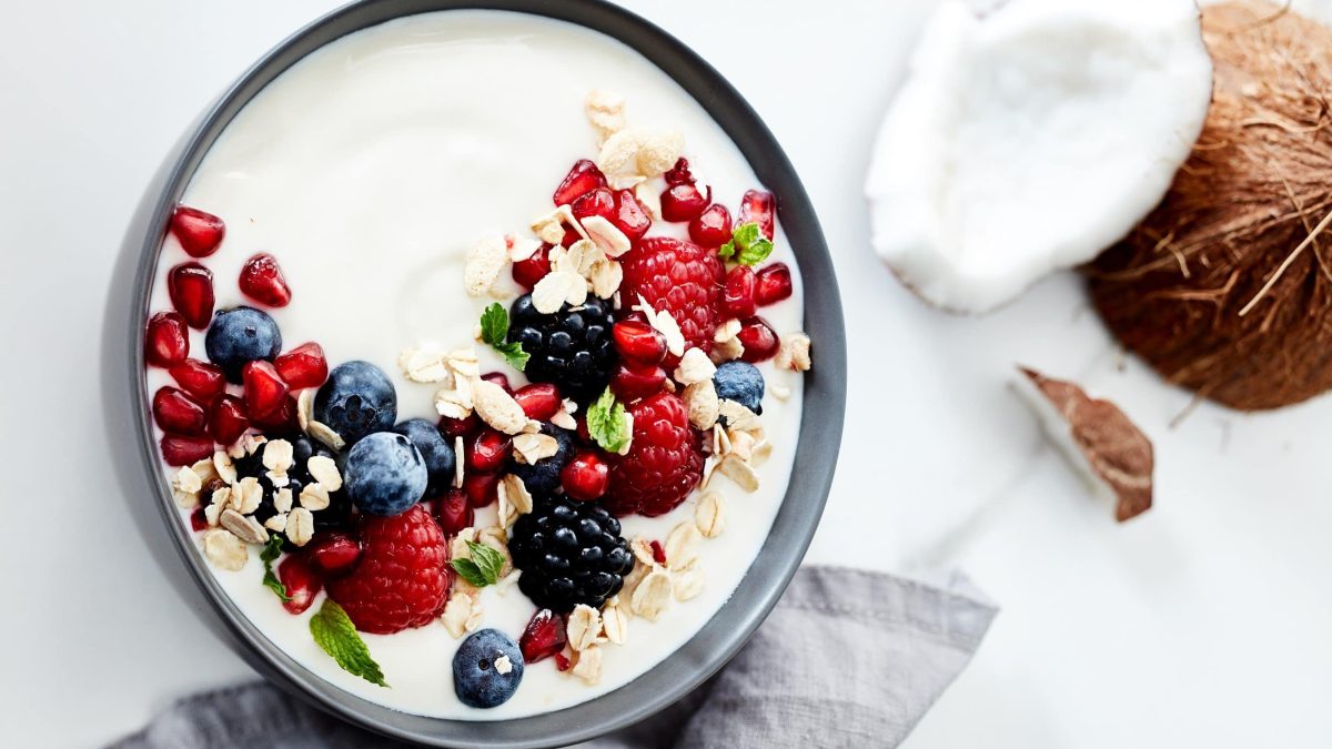 Draufsicht, heller Untergrund: graue Schüssel mit Kokos-Joghurt selber machen, darauf Beeren, Granatapfelkerne, Minze und Haferflocken, daneben Kokosnuss.
