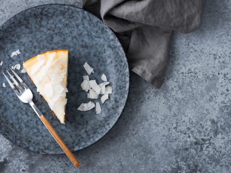 Ein blauer Tisch mit einem grauen Teller und einem Stück vom Kokosnuss-Käsekuchen mit einer Kuchengabel direkt von oben fotografiert.