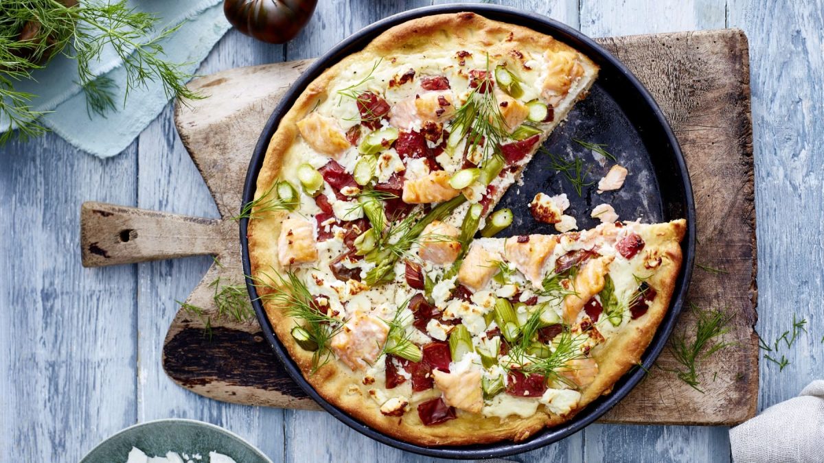 Eine Lachs-Feta-Pizza auf einer schwarzen Pizza-Backform auf einem hölzernen Schneidebrett auf einem graublau-hölzernen Hintergrund. Links oben Dill und eine Tomate auf einem hellblauen Geschirrtuch.