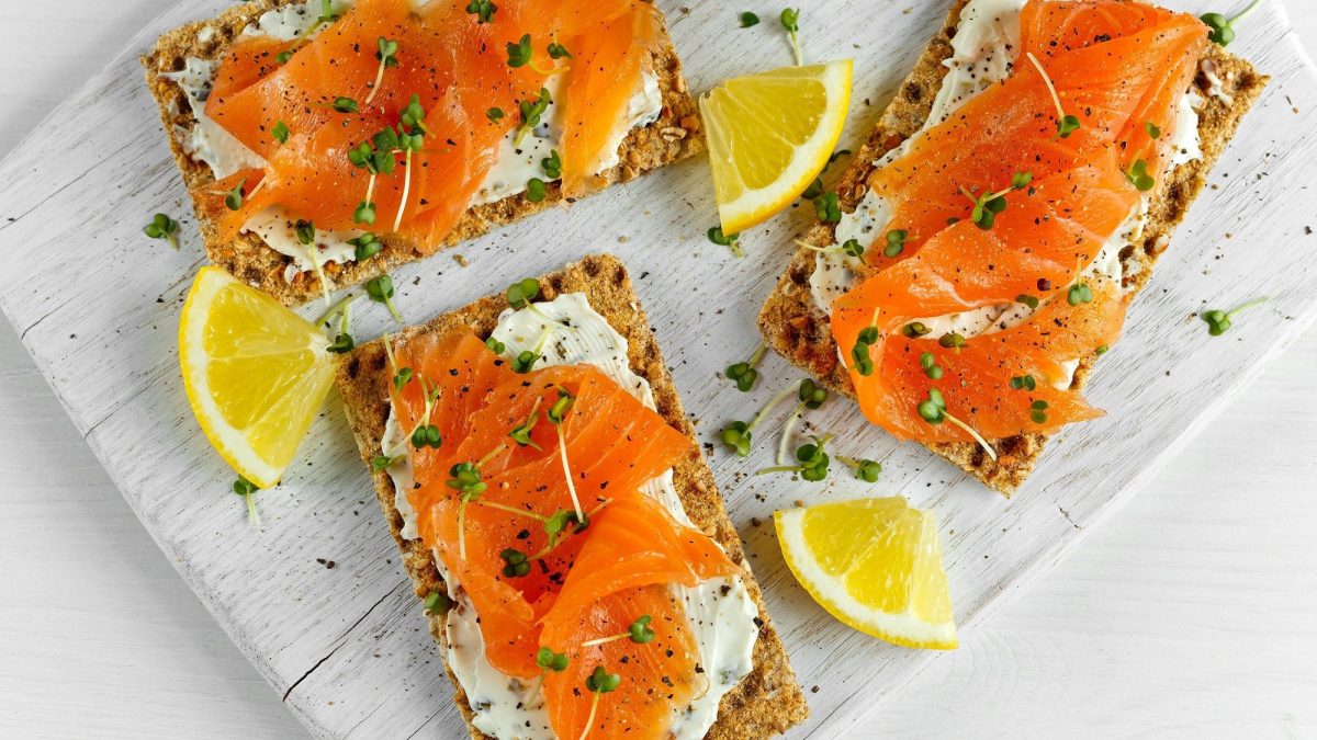 Draufsicht: heller Untergrund. Neben drei Lachs-Zitronen-Knäcke liegen kleine frische Zitronenscheiben.