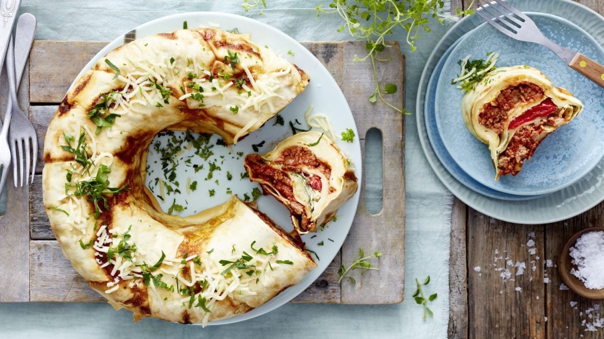 Ein weißer Teller mit einem angeschnittenen Lasagnering auf einem hölzernen Schneidebrett auf einem hellblauen Geschirrtuch auf einem hölzernen Hintergrund. Rechts ein blauer Teller mit einem Stück Lasagne, Kräuter und eine kleine Schüssel mit Salz. Links auf dem Bild drei Gabeln.