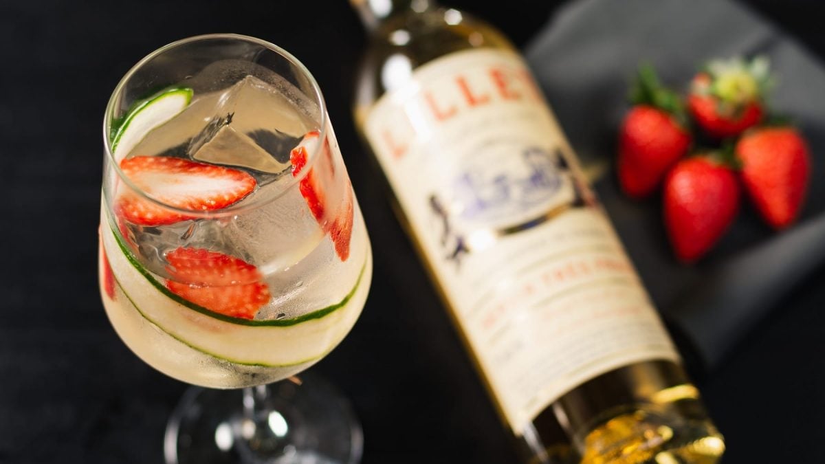 Ein Glas mit dem Drink auf schwarzem Untergrund. Rechts neben dem Glas sind unscharf ein paar Erdbeeren und eine Flasche Lillet zu erkennen.