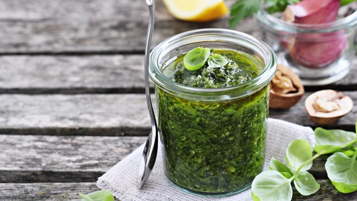 Ein Glas grünes Miso-Pesto mit Basilikumblättchen, daneben lehnt ein Löffel, drumherum Zutaten wie Walnüsse, Basilikum, Knoblauch und Zitrone.