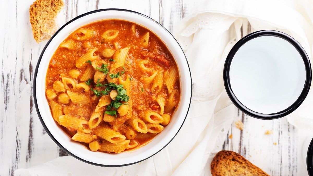 Eine Schüssel Pasta e ceci, ein italiensicher Nudeleintopf mit Kichererbsen und Tomatensuppe, in der Draufsicht, daneben ein Becher Wasser und geröstetes Brot.