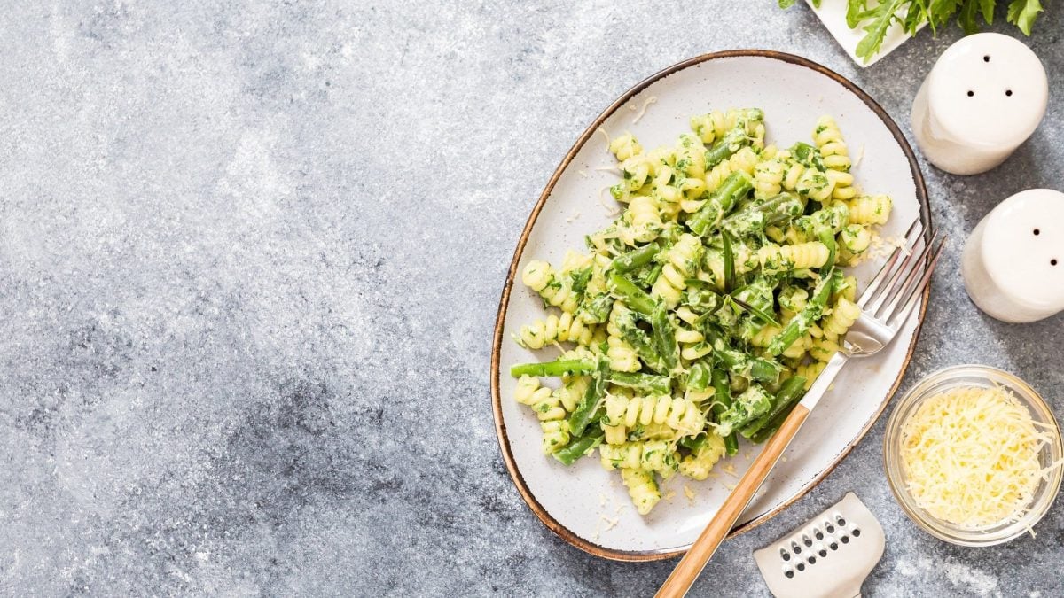 Ein Teller Pasta mit Spargel und Rucola, darauf auch eine Gabel, daneben Salz- und Pfefferstreuer, eine Schale mit geriebenem Käse, alles Draufsicht.