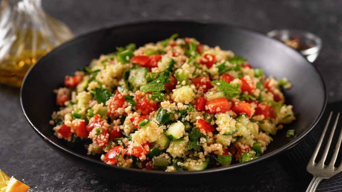 Ein Teller Pesto-Couscous-Salat mit Minze, Paprika, Gurke und Tomate, daneben eine Gabel.