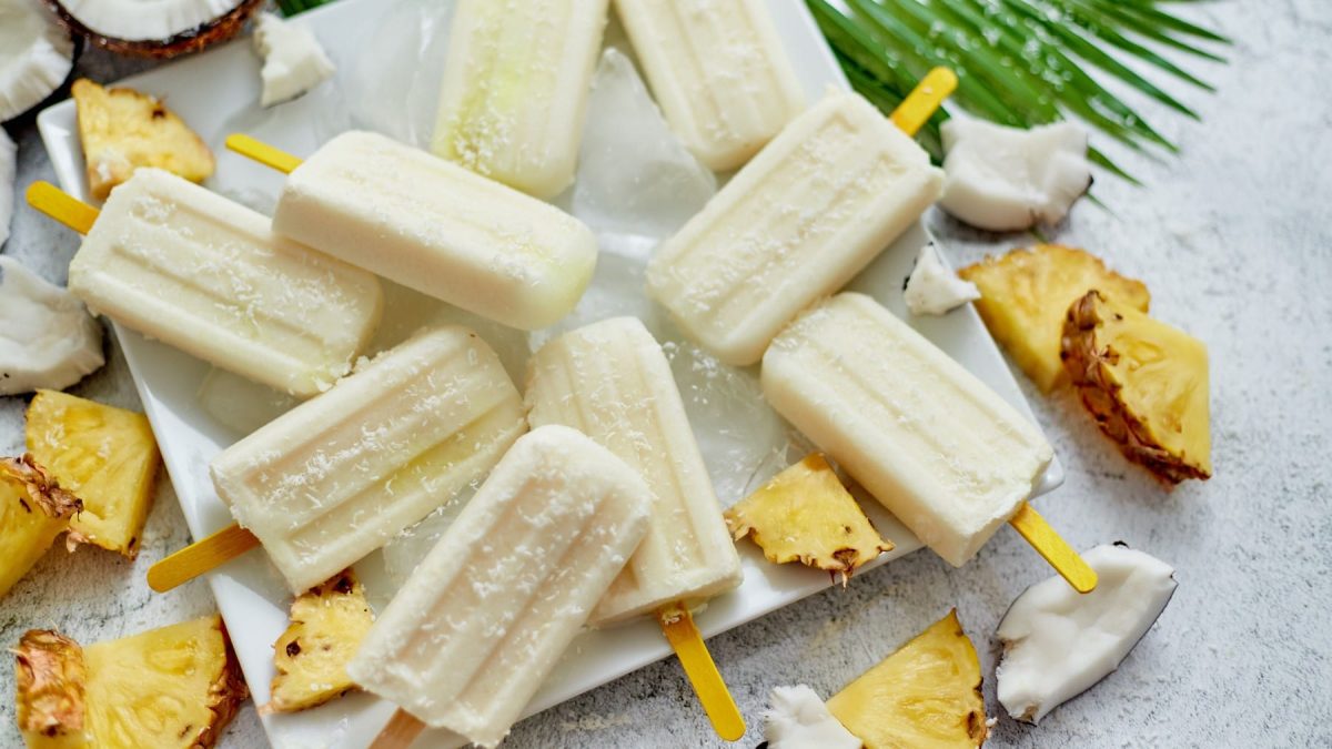 Mehrere Piña Colada Paletas auf einem weißen Teller. Drum herum Ananas- und Kokosstücke auf grauem untergrund.