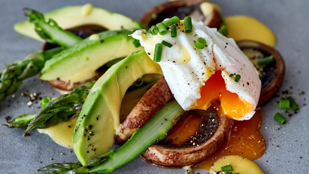 Nahaufnahme von Pilzen, grüner Spargel, pochiertes Ei, Avocado und Schnittlauch auf einem grauen Teller.