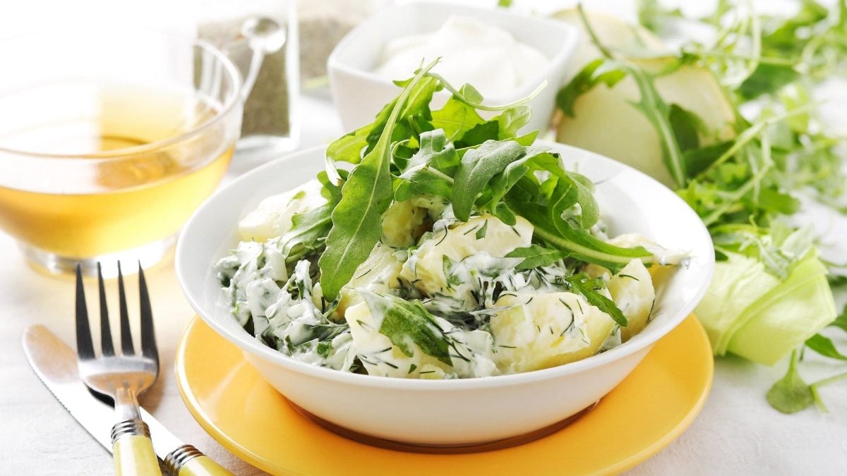 Eine Schale Rucola-Kartoffelsalat mit griechischem Joghurt und Dill, daneben eine Gabel und Zutaten.