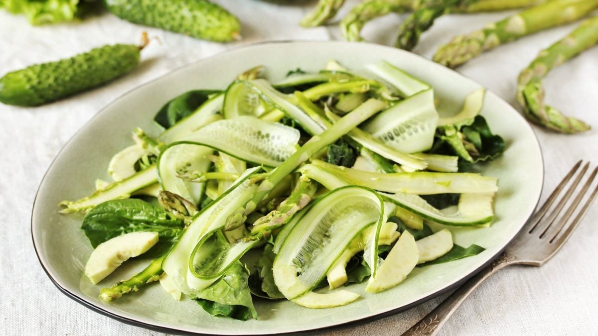 Ein Teller Spargel-Gurken-Salat, mit Babyspinat, daneben eine Gabel und Gurken sowie Spargelstangen.