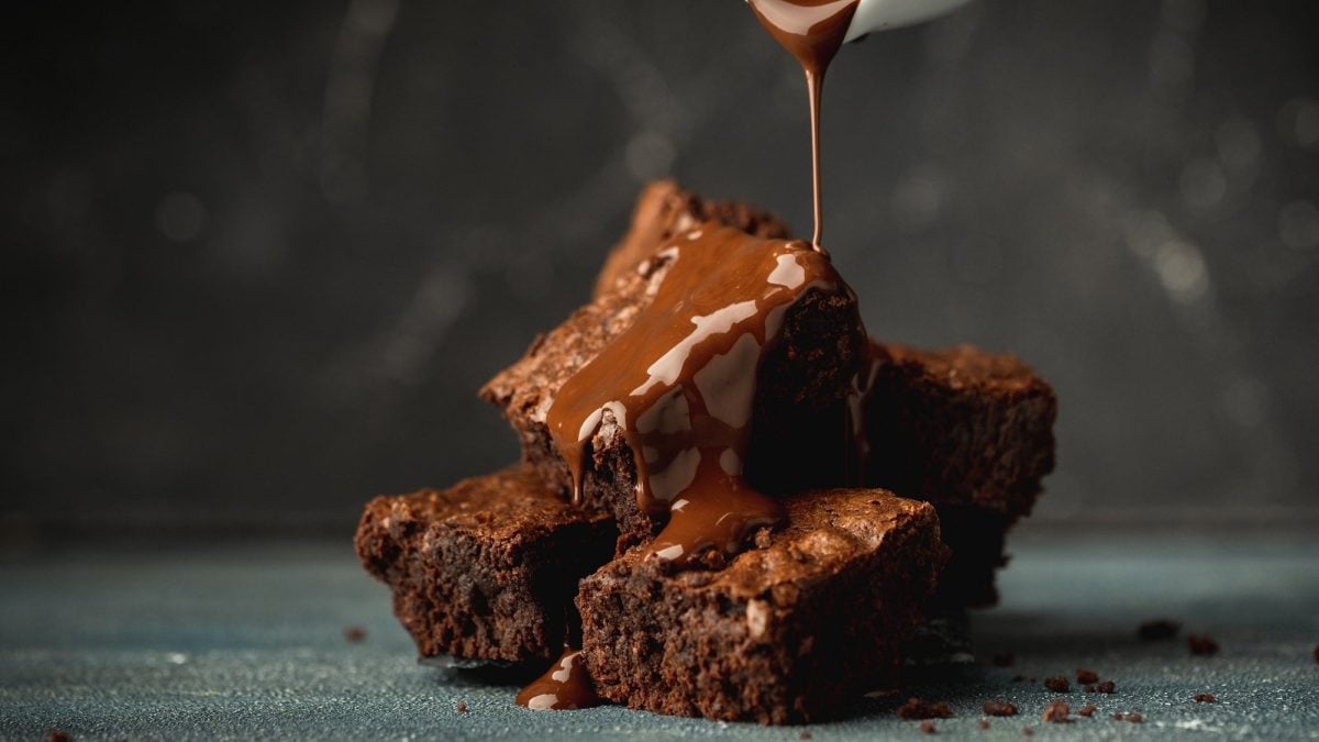 Ein paar Tahini-Brownies auf ebenem Untergrund, die mit flüssiger Schokolade übergossen werden.