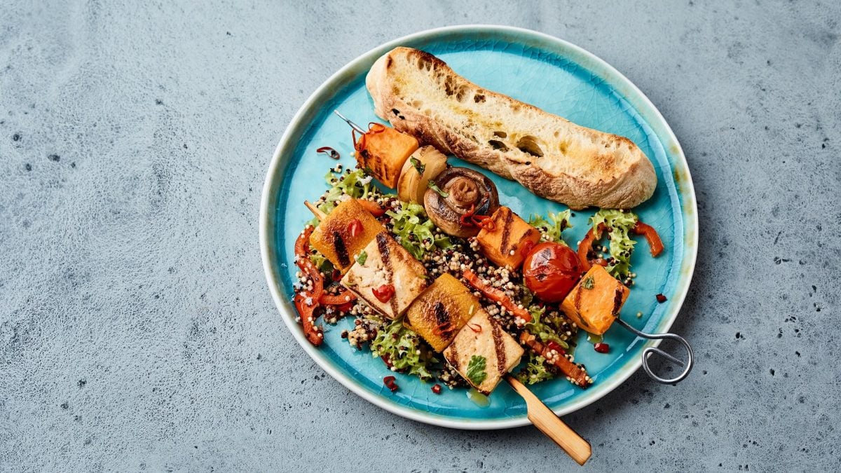 Zwei gegrillte Tofu-Gemüsespieße auf einem Teller, daneben eine Scheibe Baguette.