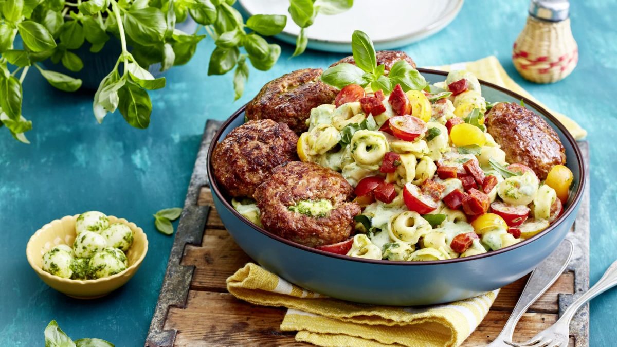 Eine blaue Bowl mit Tortellinisalat und Hackbällchen auf einem gelben Geschirrtuch auf einem hölzernen Brett. Der Hintergrund ist blau und links eine kleine Schüssel mit Mozzarellakugeln und ein Basilikumstrauch. Rechts oben ein Salzstreuer.